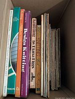 Photo of a box filled with various knitting and crochet books and pattern booklets, showing spines with titles related to knitting, afghan, and baby clothes patterns.
