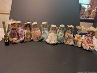 Full lineup of 10 porcelain collectible dolls dressed in vintage attire varying in colors and styles, seated and standing against a dark background.