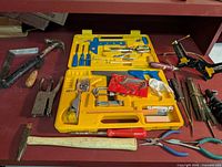 View of assortment of hand tools on a red surface, including yellow tool kit with various tools, stapler, hammers, pliers, files, screwdriver and glue gun.