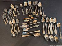 Full layout of the mixture of silver teaspoons and dessert forks showing varied designs and some tarnish.