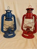 Two vintage style oil lamps side by side on fabric backdrop, one blue and one red, showing overall size and condition