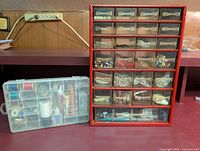 Red multi-drawer organizer filled with various sewing notions, buttons, hooks, pins, and thread spools next to a plastic container with sewing threads and glues.