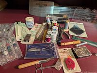 Photo showing a variety of sewing notions including thread spools, scissors, pincushion, measuring tapes, needle packets, and sewing machine accessories on a surface with a green cutting mat in the background.
