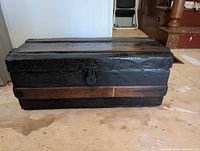 Front view of the dark wooden trunk showing the latch lock and raised wooden slats on top, placed on a wooden floor.