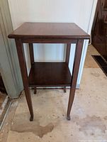 Front view of tall wood side table showing square top, lower shelf, and slender legs.