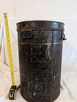 Front view of tall cylindrical black metal tin storage container with visible latch and side handle, scale next to it showing about 22 inches height.