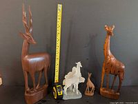 Seven wood animal sculptures arranged on a black surface with a yellow measuring tape for scale, showing a large antelope and giraffes height over 12 inches.