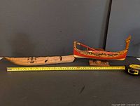 Two wooden boat models side by side on a black surface with a tape measure for scale, showing the natural canoe and the colorful Maltese boat