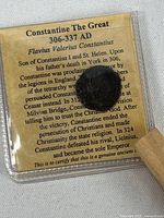 Ancient dark coin housed in a clear plastic holder along with a historical document about Constantine the Great in the background.
