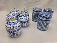 Five blue and white Chinese porcelain jars with lids arranged in two rows, showing variety of designs with calligraphy and figurine toppers.