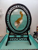 Front side of the oval silk screen showing an orange and white cat playing with a cricket, mounted on carved wooden stand