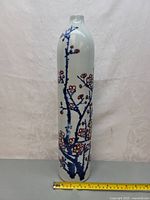 Full view of tall slender porcelain vase showing blue branches and red flowers painted on white background with measuring tape at base.