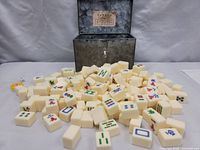 Mahjong tiles scattered with the storage case behind them, showing various tile faces, colors, and the open storage box with label inside the lid.