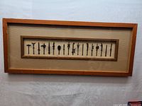 Framed artwork featuring 18 miniature bronze replicas of ancient Chinese weapons displayed in a wooden frame with beige background.
