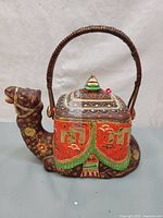 Front view of the camel-shaped teapot with detailed moriage and colorful painted patterns, wicker handle attached.