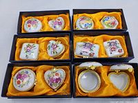 Full set of 6 vintage Chinese porcelain trinket boxes displayed in black boxes with yellow satin lining. Various floral painted patterns and shapes visible including round, heart, rectangular, and oval shapes.