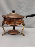 Full view of the copper pot with lid, black handle, and brass legs on the heating base.