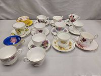 Set of fourteen vintage tea cups and saucers arranged in rows on a gray surface showing various floral patterns and gold trim.