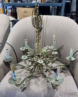 Front view of antique brass chandelier showing six arms with floral leaf decorations and candle-style light sockets, placed on a beige upholstered chair.