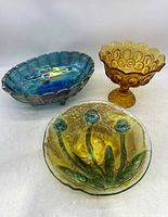 Three collectible glass pieces: amber compote on the right, blue carnival glass oval bowl on the left, and floral art glass bowl in front.