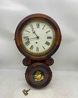 Front view of the antique wall clock showing the clock face with Roman numerals, wooden frame, and winding key on the surface.