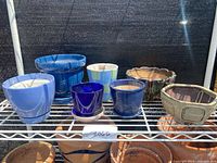 Overview of all seven flower pots on a white metal shelf against black mesh background, showing different shapes, sizes, and colors.