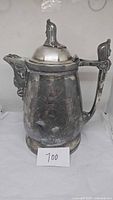 Front side view of large vintage silver-tone water pitcher showing ornate lid with head figure and decorative body pattern, standing about 13 inches tall.