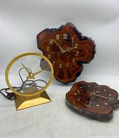 Three clocks on white surface. One Jefferson Golden Hour electric clock with gold metal frame and clear face, unpowered and untested. Two live edge wood clocks with shellacked finish, one with Arabic numerals, one with Roman numerals.