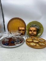 Overview photo showing comedy and tragedy copper tone wall hangings, woven rattan tray, green ceramic plate, wooden tray with ceramic coasters, etched blue stem and leaf pattern coupe glasses, and small silver candle holder.