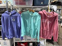 Three hooded sweatshirts on hangers in purple, teal, and pink colors, displayed hanging on a rack.