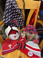 Collection of Phillies winter hats including red knit hat, two trapper hats, gray beanie, one pair of utility gloves, and a black Phillies travel bag with repeated logo pattern.