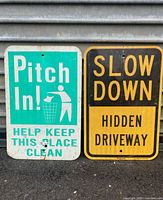 Two metal street signs side by side: one teal 'Pitch In!' sign and one black and yellow 'Slow Down' sign.