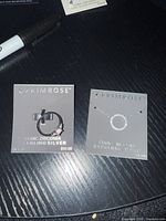 Two items on black surface: a ring on grey card with cubic zirconia stone and retail price $50, and a circle pendant necklace on grey card with retail price $45.