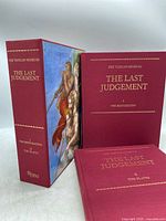 Two hardcover books in red covers with gold embossed titles 'The Last Judgement I The Restoration' and 'The Last Judgement II The Plates' next to a colorful slipcase with artwork from The Last Judgement fresco.