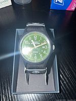 Front-facing image of Timex Expedition watch showing green dial with white numbering and date, placed in black box on dark surface.