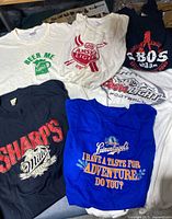 Photo showing the six t-shirts arranged together displaying logos and colors including white, blue, and black shirts with various beer brand logos.
