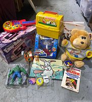 Photo showing boxed Easy-Bake Oven, yellow Snoopy lunchbox, M&M candy dispenser, plush teddy bear, Hulk action figure in packaging, Rubik's Cube, and several smaller toys including figurines and spinning top.