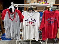 Photo showing three women's Phillies shirts hanging on hangers: gray/red hoodie, white V-neck, red V-neck.