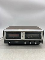 Front and top view of a vintage Zenith alarm clock radio with woodgrain top, metallic front panel, analog radio tuner, digital clock display, and control knobs.
