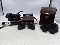 Three pairs of black binoculars with original brown leather cases shown side by side on white background.