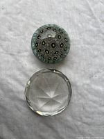 Top view of abstract multicolor glass paperweight and clear diamond-faceted paperweight on white fabric.
