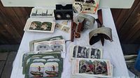 Wide shot of the collection showing multiple stereo view cards spread out and stacked, two vintage stereo viewers including a black plastic viewer and a gray metal and leather viewer, plus some accessories on a white table.