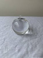Front view of crystal apple paperweight showing the transparent clear crystal and small stem on top. Displayed on white fabric background.