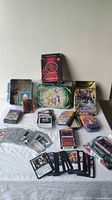 Full lot display including multiple loose cards, booster packs, and metal storage tins featuring Pokemon and Yu-Gi-Oh artwork.