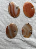 Four oval orange agate stone pendants arranged on light fabric background showing their polished surfaces and banded patterns with drilled holes.