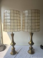 Two vintage brass lamps with tall, curved bases and capiz shell shades in a grid pattern, placed side by side on a white cloth.