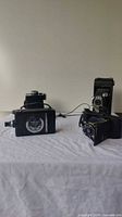 Photo showing five vintage cameras arranged on a white surface with different designs including box and bellows style
