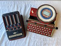 Blue and red mechanical adding machine and red Simplex typewriter side by side on a cloth surface.