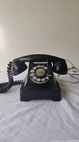 Front view of entire black Bakelite rotary dial telephone with handset, resting on a white surface.