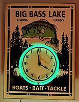 Front view of Big Bass Lake neon clock illuminated showing green neon light around clock face and fishing cabin themed graphics with large bass fish.